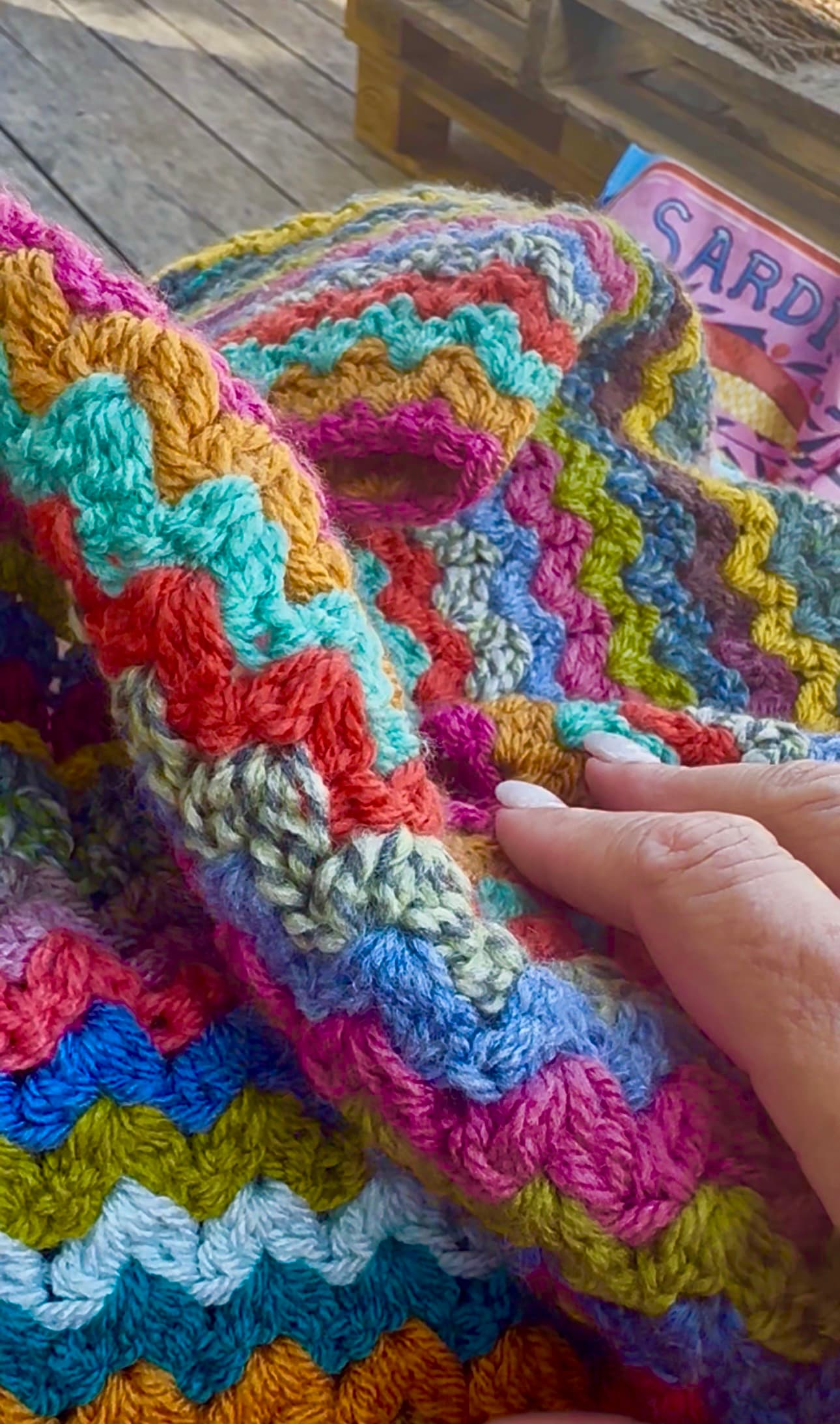 A hand resting on a chunky, rainbow-striped ripple crochet blanket on a sunny wooden deck, with a pink sardine tin nearby.